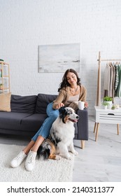 Happy Young Woman Sitting On Couch And Cuddling Australian Shepherd Dog While Holding Cup Of Coffee