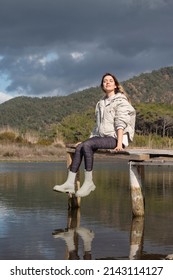 Happy Young Woman Sitting On Old Small Dock. Closed Eyes. Beautiful Landscape View. Nature Admiration. Dangling Legs From Jetty. She Is Sitting On Wooden Dock Above Sea. 