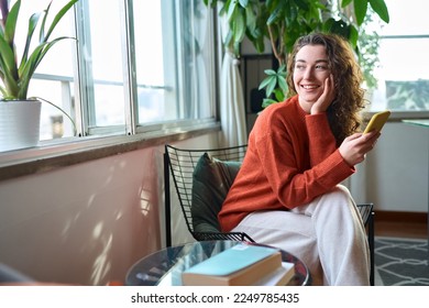 Happy young woman sitting on chair holding mobile phone using cellphone device, checking modern apps on smartphone, texting messages, browsing internet doing shopping relaxing at home.