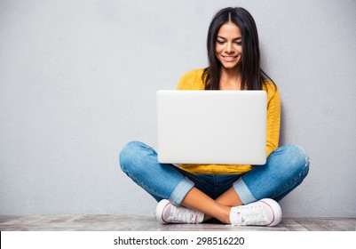 Happy young woman sitting on the floor with crossed legs and using laptop on gray background
