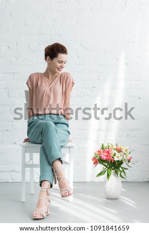 happy young woman sitting on chair with flowers on floor in front of white brick wall