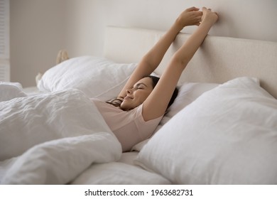 Happy Young Woman Sleeping In Comfortable Bed On Orthopedic Mattress. Peaceful Teen Girl Awaking In Bedroom Early In Morning, Stretching Arms And Body, Smiling With Closed Eyes, Enjoying Good Bedtime