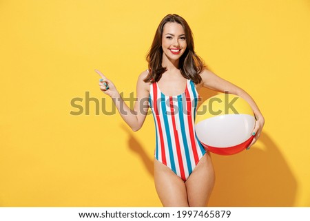 Happy young woman slim body wear striped red blue one-piece swimsuit hold play inflatable beach ball isolated on vivid yellow color wall background studio. Summer hotel pool sea rest sun tan concept