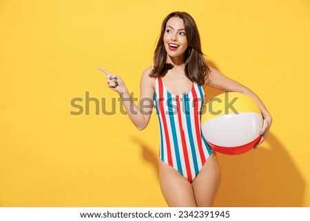 Happy young woman slim body wear striped red blue one-piece swimsuit hold play inflatable beach ball isolated on vivid yellow color wall background studio. Summer hotel pool sea rest sun tan concept