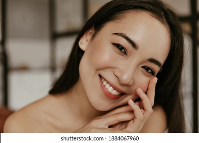 Happy Young Woman Smiles Sincerely. Attractive Brunette Asian Lady Touches Gently Her Face Skin And Looks Into Camera In Good Mood.