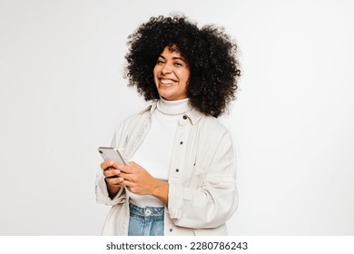 Happy young woman smiling at the camera while holding her smartphone. Cheerful woman with curly hair sending a text message while standing against a white background. - Powered by Shutterstock