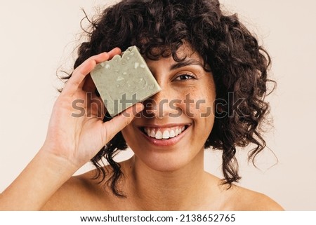Happy young woman smiling at the camera while holding a bar of natural soap close to her face. Beautiful young woman using handmade cosmetic soap to take care of her skin.