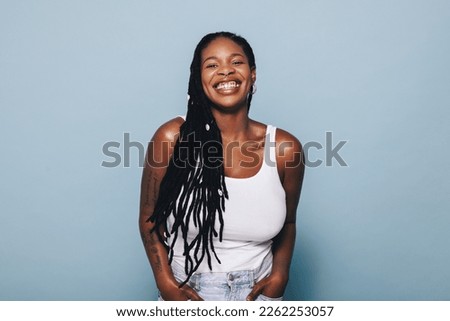 Happy young woman smiling at the camera while standing against a blue background in casual clothing. Stylish young black woman wearing arm tattoos and piercing jewellery confidently in a studio.