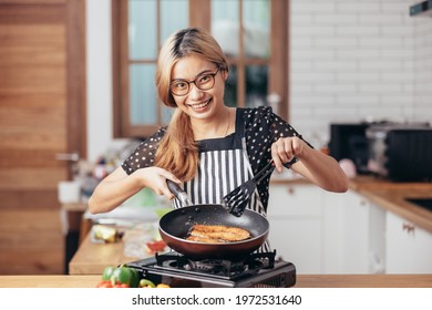 Happy Young Woman Smiling In Modern Kitchen. Beautiful Asian Woman Stands In A Kitchen Cooking Wearing A Red Apron. The Healthy Menu She Will Cook Is Salmon, Steak And Salad. Modern Woman Kitchen