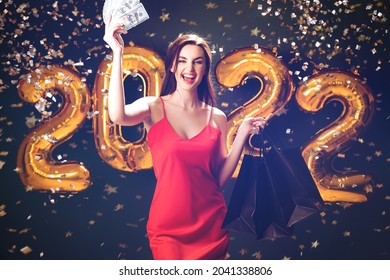 Happy young woman smiling in red dress raises dollars and purchases up on black background with golden numbers 2022 balloons confetti. New Year shopping. Black Friday. Celebrate sales .