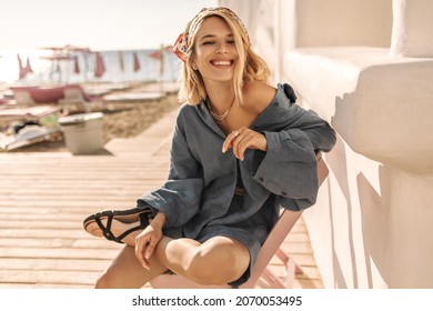 Happy young woman with snow-white cute smile sits on beach. Light-skinned short-haired blonde with bandana on her head in blue summer clothes. Concept of enjoying weekend, vacation - Powered by Shutterstock