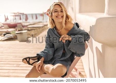 Happy young woman with snow-white cute smile sits on beach. Light-skinned short-haired blonde with bandana on her head in blue summer clothes. Concept of enjoying weekend, vacation