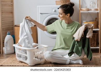 Happy young woman sorting clothes near washing machine in laundry room - Shutterstock ID 2265425303