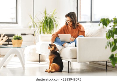 Happy young woman spending time with her pet beloved dog at home sitting on couch and browsing news in  tablet - Powered by Shutterstock