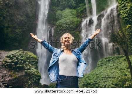 Happy young woman spreading hands enjoying nature with waterfall in background. Caucasian female standing in front of a waterfall with her arms outstretched.