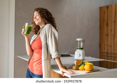 Happy young woman standing in kitchen and holding glass of detox juice. Cheerful girl drinking healthy smoothie at home. Beautiful smiling woman drinking green vegetable smoothie. - Shutterstock ID 1929927878
