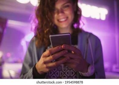 Happy Young Woman Standing On Festively Lit Purple Neon Signs City Street At Night, Typing On Mobile Phone And Smiling.