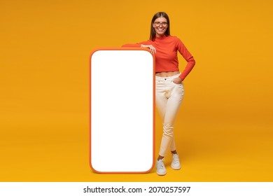 Happy young woman standing and showing blank screen of huge phone, mockup for your app, isolated on yellow background