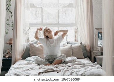 Happy Young Woman Stretching In Bed Waking Up In Cozy Bedroom With Bohemian Interior Style. Female Wearing In Comfort Pajamas Enjoying Early Morning, Resting Alone