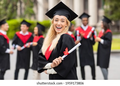 Happy Young Woman Student Cheerfully Posing While Graduation Party, Wearing Graduation Robe And Hat, Holding Diploma And Smiling At Camera, International Group Of Students On Background