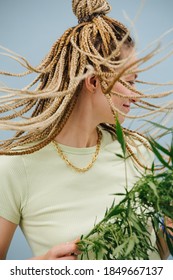 Happy young woman with stylish blond afro braids over blue background. Waving her hair, holding cannabis plant in a hand.