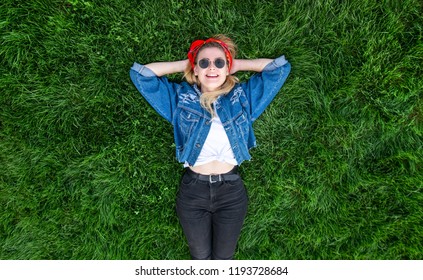Happy young woman in stylish clothes lays on a green, beautiful lawn, looks at the camera and smiles. Top view. Attractive lady lies on the grass. Copyspace