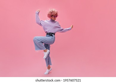 Happy young woman in stylish jeans and purple sweater dance on pink background. Curly short-haired girl move in great mood on isolated background .