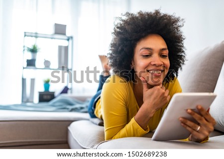 Happy young woman with tablet pc laying on sofa. Young happy woman lie down on bed and playing smart tablet at home. Beautiful young black women using tablet computer