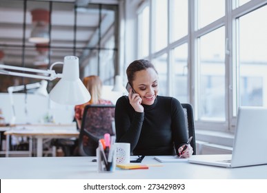 Happy young woman taking notes while talking on mobile phone. African woman working at her desk answering a phone call.