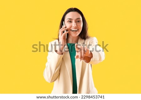Happy young woman talking by phone and pointing at viewer on yellow background
