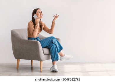 Happy young woman talking on smartphone, sitting in armchair against white studio wall, empty space. Cheerful millennial lady making phone call, having conversation, enjoying communication - Shutterstock ID 2049512792