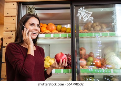 Happy Young Woman Talking On Cell Phone And Buying Pomegranate At Grocery Store