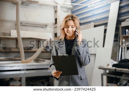 Happy young woman talking on phone at carpentry production of DIY wooden furniture. Concept call center or owner small business.