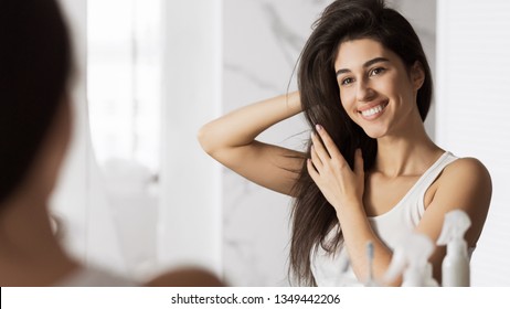 Happy young woman touching her hair and smiling looking in mirror. Home haircare concept