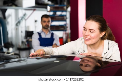 Happy Young Woman Touching Surface Of Repainted Car Body In Auto Repair Shop