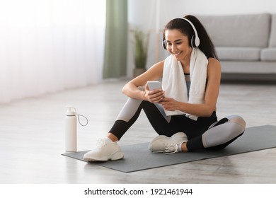 Happy young woman with towel on shoulders and wireless headset sitting on yoga mat in living room, listening to music while resting, copy space. Sports music mobile application concept - Powered by Shutterstock