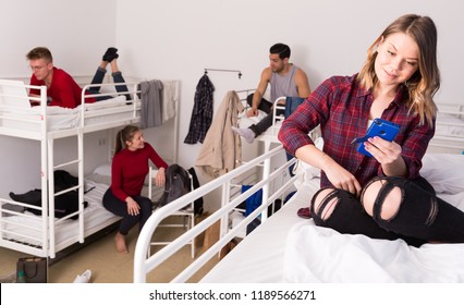 Happy Young Woman Traveler Resting With Phone On Two-story Bed In Hostel Dorm