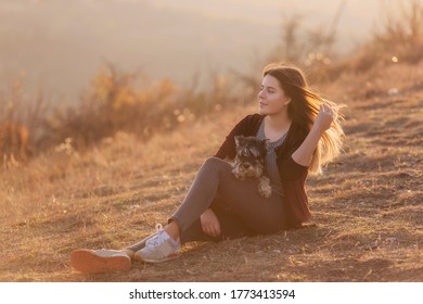 Happy Young Woman Travels With Her Domestic Dog Black Schnauzer. A Beautiful Girl Hugs A Pet, Sits On A Mountain, The Wind Develops Her Red Long Hair, She Smiles. Vacation Concept With Pets. Caring 