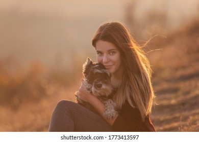 Happy Young Woman Travels With Her Domestic Dog Black Schnauzer. A Beautiful Girl Hugs A Pet, Sits On A Mountain, The Wind Develops Her Red Long Hair, She Smiles. Vacation Concept With Pets. Caring 