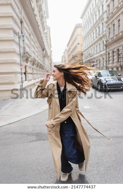 happy young woman in trench coat looking away\
while walking on street in windy\
day