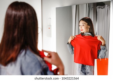 Happy young woman trying on clothes in dressing room and looking at the mirror while shopping in fashion store