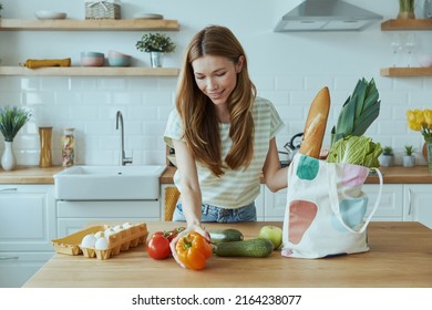 Happy young woman unpacking the bag with healthy food while standing at the domestic kitchen - Powered by Shutterstock