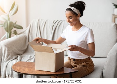 Happy Young Woman Unpacking Delivery Box, Sitting On Couch By Tea Table, Home Interior. Satisfied Customer Cheerful Lady Opening Package, Ordering Clothes Or Cosmetics On Internet, Copy Space