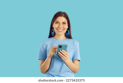 Happy young woman uses mobile phone for online shopping, chatting, using mobile apps or browsing social networks. Smiling woman in casual t-shirt holding cellphone in hands on light blue background. - Shutterstock ID 2273433375