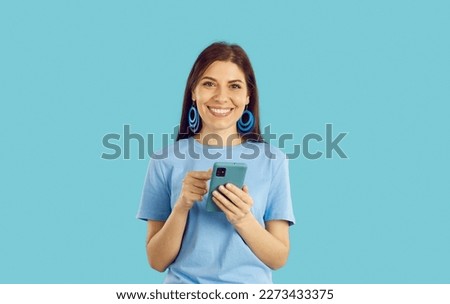 Happy young woman uses mobile phone for online shopping, chatting, using mobile apps or browsing social networks. Smiling woman in casual t-shirt holding cellphone in hands on light blue background.