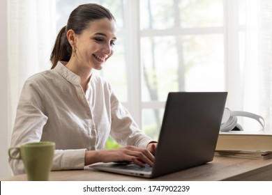 Happy Young Woman Using Her Laptop At Home, She Is Sitting At Desk And Smiling, Window With Beautiful View In The Background