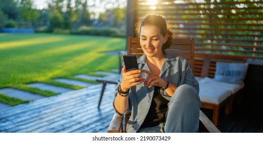 Happy Young Woman Is Using A Phone Sitting On The Patio In Summer. 