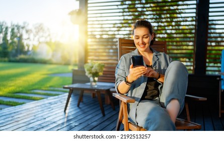 Happy Young Woman Is Using A Phone Sitting On The Patio In Summer. 