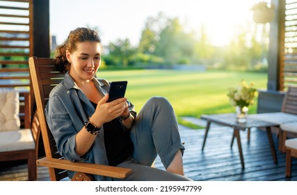 Happy Young Woman Is Using A Phone Sitting On The Patio In Summer. 