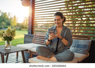 Happy Young Woman Is Using A Phone Sitting On The Patio In Summer. 